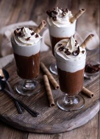 3 Chocolate Mousse Parfaits with spoons, whipped cream and chocolate shavings on a wooden board